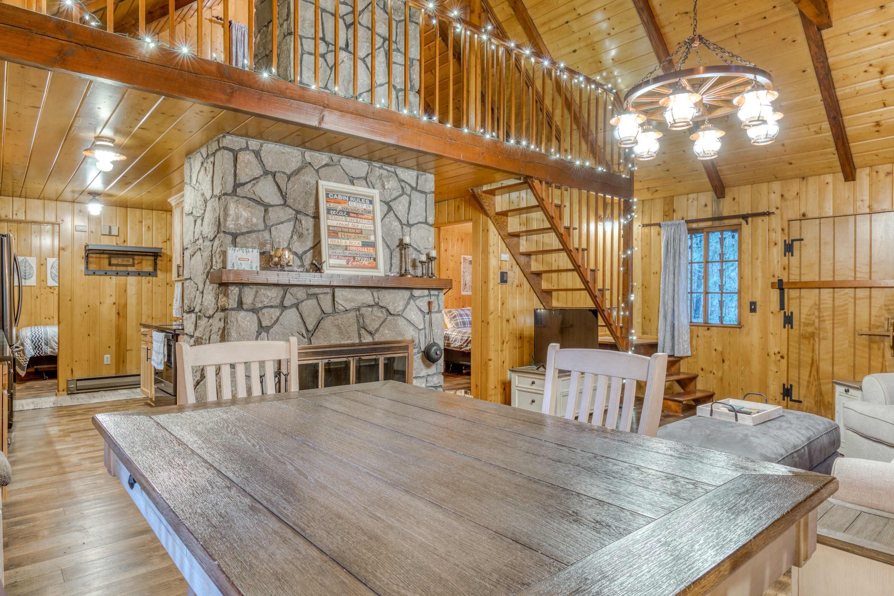 Interior de una cabaña de madera con chimenea de piedra, una mesa de comedor y una escalera.