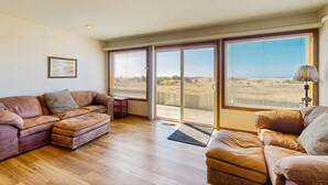 Una sala de estar con sofá, reposapiés y una pequeña mesa. Hay un ventanal con persianas, una puerta que da al exterior y una lámpara sobre una mesita.
