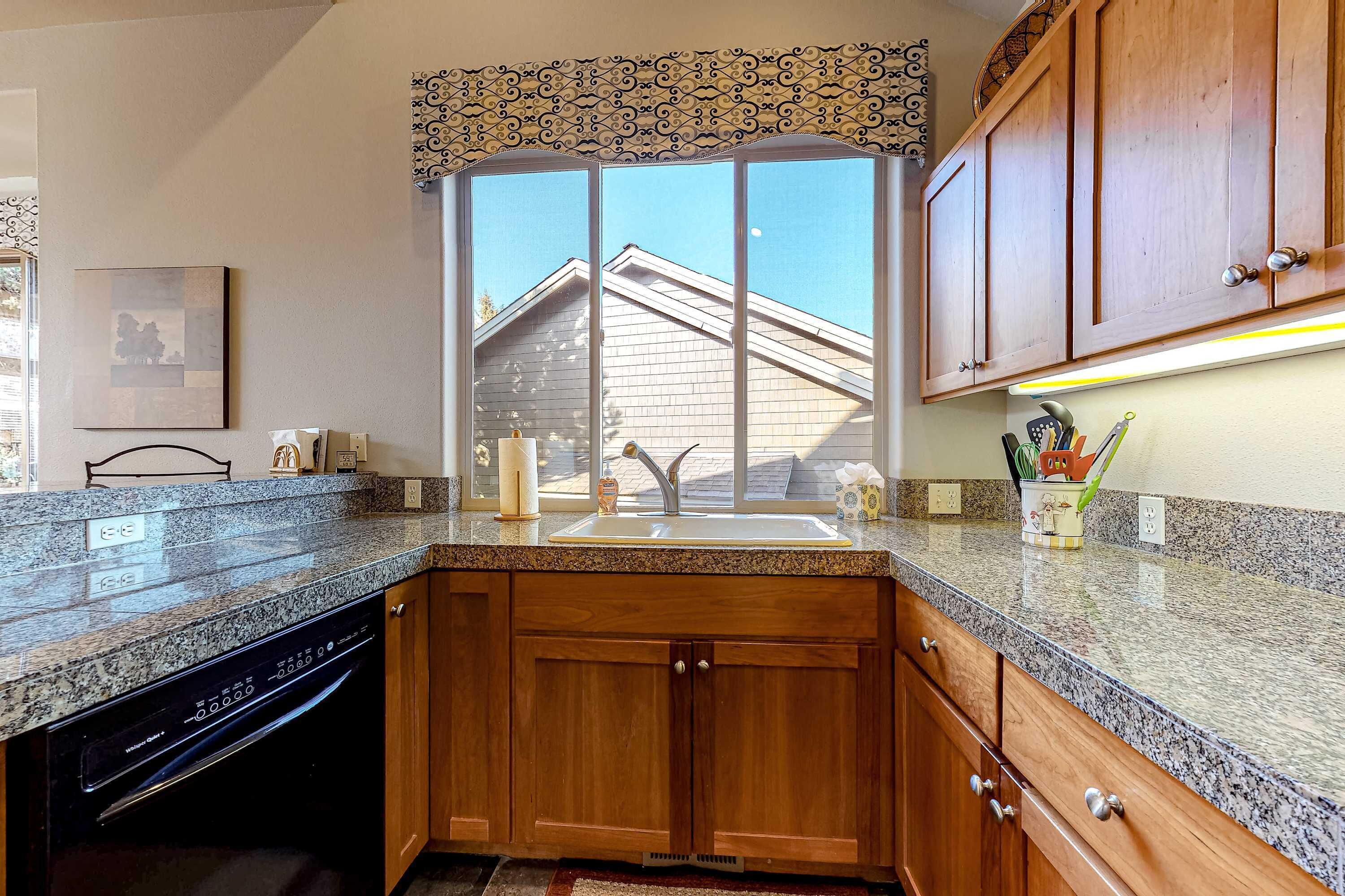 A kitchen with wooden cabinets, a granite countertop, a built-in dishwasher, and a window with a view of a house.