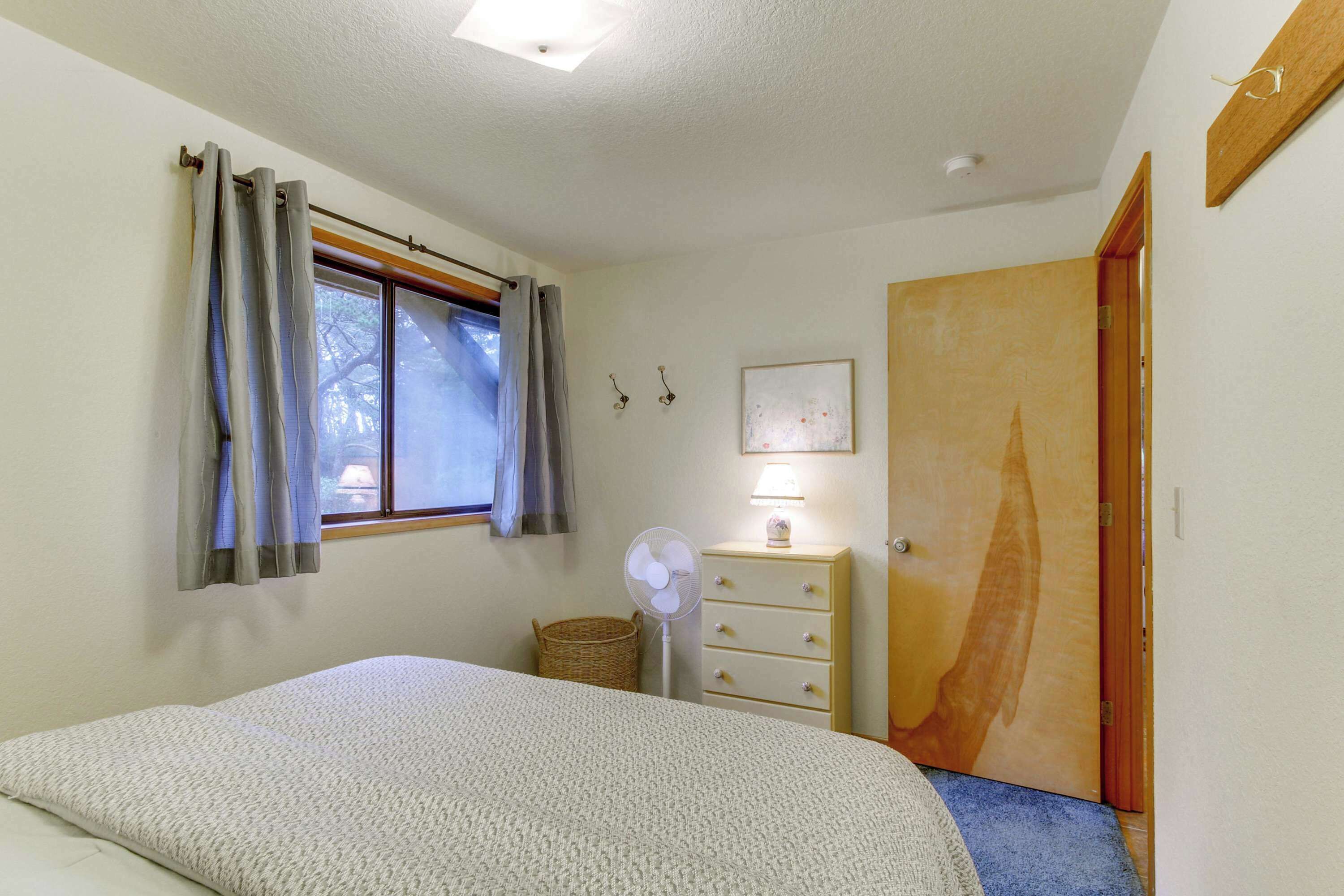 A bedroom with a bed, a window with curtains, a bedside lamp, a fan, a wooden door, and a wall-mounted picture.