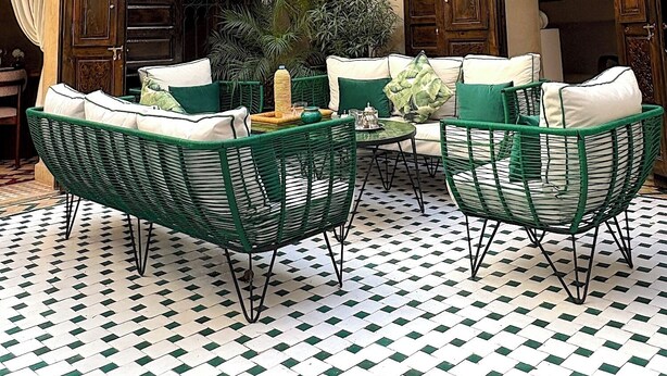 Een terras buiten met een geruit vloertje, groen rieten meubilair en een sierhekwerk.