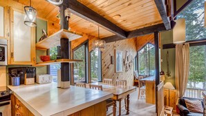 Interno di una baita in legno con angolo cottura, zona pranzo e soggiorno, caratterizzato da ampie finestre e vista sull'esterno.