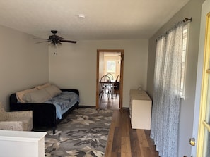 A room with a bed, a ceiling fan, a patterned rug, a dining area with chairs and a table, and a door with patterned curtains.