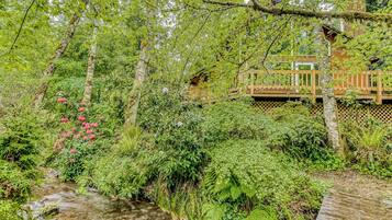 Une terrasse en bois, entourée d’une clôture en bois, et bordée d’un feuillage verdoyant et dense ainsi que d’un petit ruisseau.