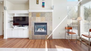 A modern living room with a stone fireplace, white cabinetry, a flat-screen TV, and a large window with blinds. - Spacious townhouse with fireplace, fast WiFi & resort pools-hot tub-golf (Eagle Crest)