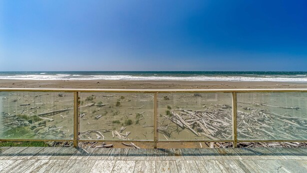 Blick auf einen Strand von einer Holzterrasse mit Glasgeländer, Ozeanwellen und Treibholz am Ufer.