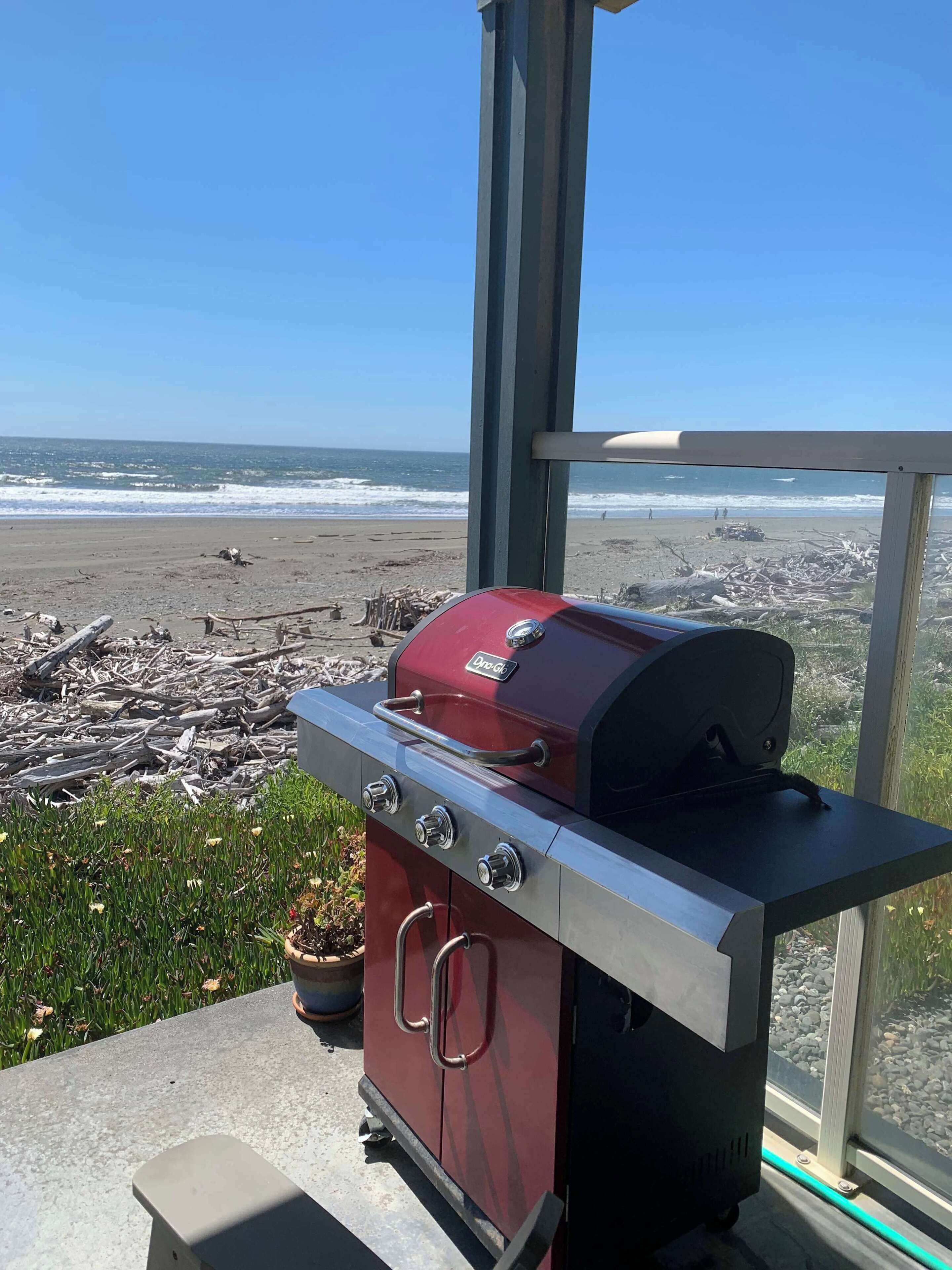 Un barbecue rouge pour l’extérieur, doté de quatre brûleurs, placé sur une surface en béton et offrant une vue sur la plage et l’océan.