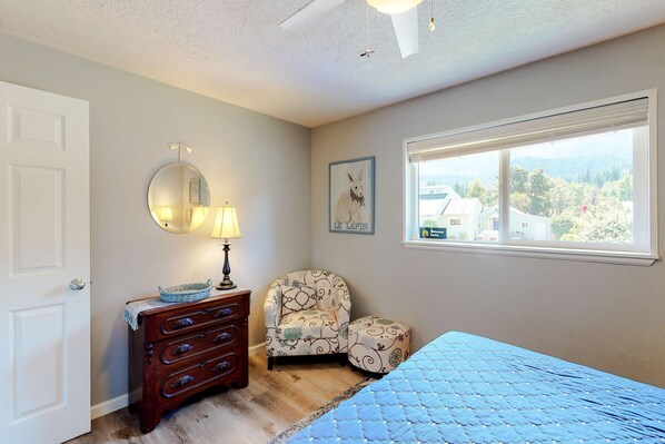 A bedroom with a bed, a dresser, a chair, a window with a view, and a ceiling fan.