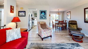 Un salón amplio con un sofá rojo, un sillón estampado y un mecedora. Se ve un comedor con mesa y sillas al fondo.