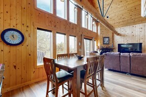 A wooden interior with a dining table, chairs, a sofa, a TV, and large windows. - Beautifully updated, golf-course-front chalet with deck, pools, hot tubs & gyms (Eagle Crest)