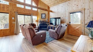Ein Holzhütten-Innenraum mit Kamin, Ledersesseln, einem Fernseher und großen Fenstern.