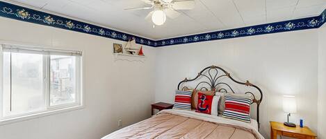 A bedroom with a bed, a nightstand, a ceiling fan, a window, and a decorative border on the wall.