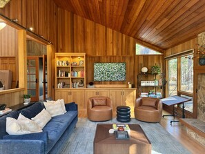 Un vaste espace de vie comprenant un canapé bleu, des fauteuils en cuir, une table basse, des étagères en bois, un téléviseur et un mur en pierre.
