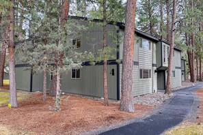 A two-story building with a grey exterior, surrounded by tall pine trees, and a paved driveway leading to the entrance.
