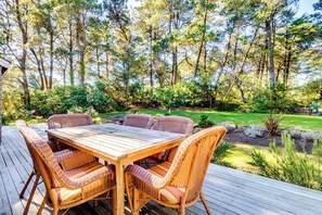 Une terrasse en bois avec une table et des chaises en osier, entourée d’un jardin et d’arbres.