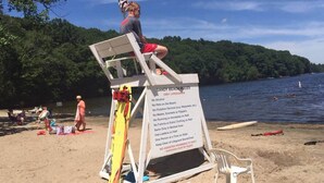 Ein Rettungsschwimmerstand mit einer Übersicht über Strandregeln an einem sandigen Strand, im Hintergrund ein See.