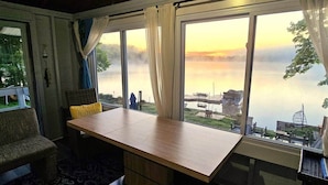 Ein Zimmer mit Tisch und Stühlen, großen Fenstern mit Vorhängen und Blick auf ein Gewässer.