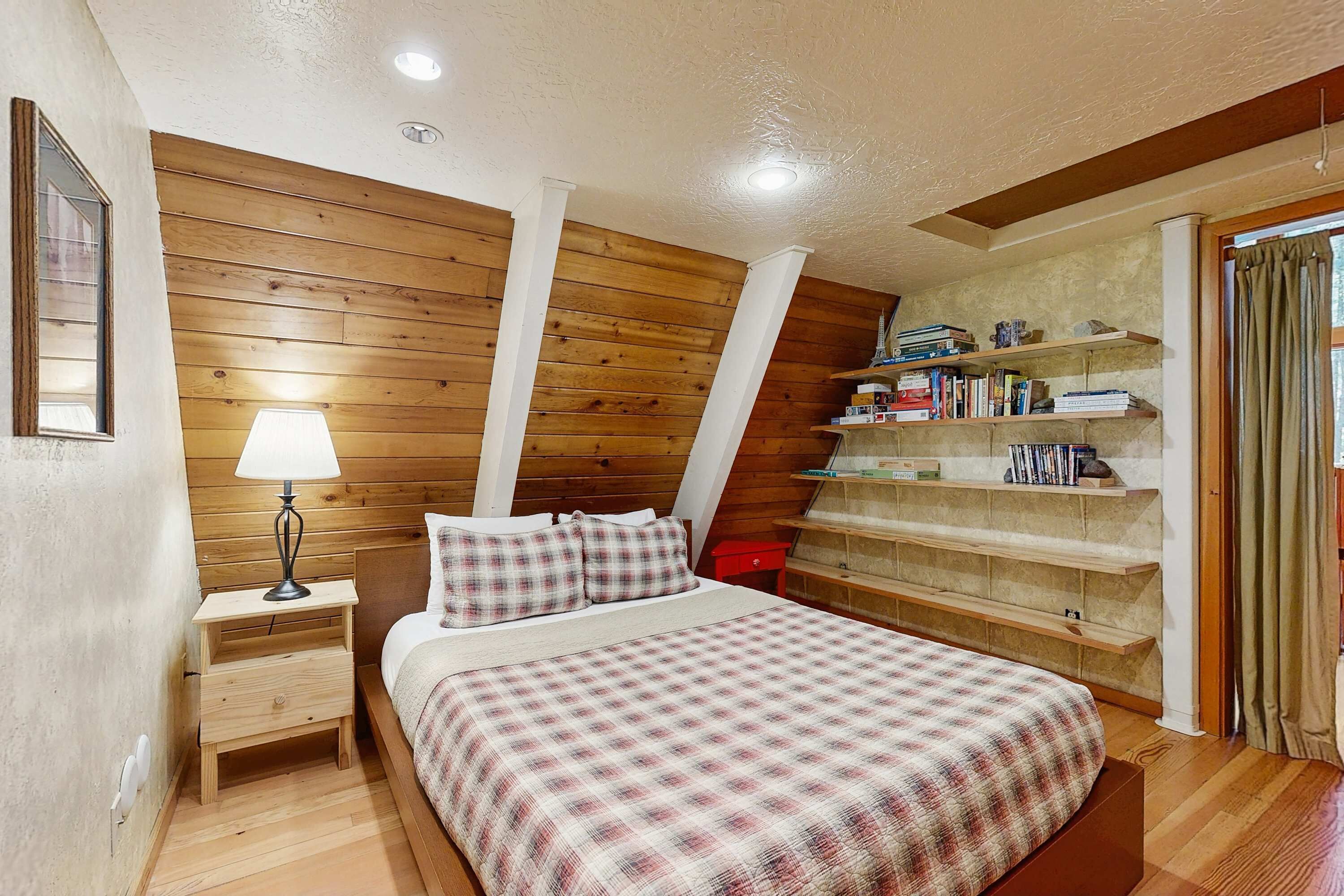 A bedroom with a wooden sloped ceiling, a bed with a plaid bedspread, a nightstand with a lamp, and built-in shelves with books and decorative items.