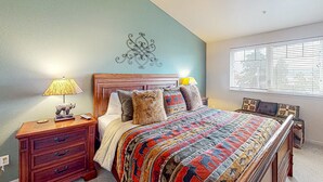 A bedroom with a wooden bed, a nightstand with a lamp, a decorative wall piece, and a window with blinds. - Classy and Convenient Dog-Friendly Condo with Private Balcony (Florence)
