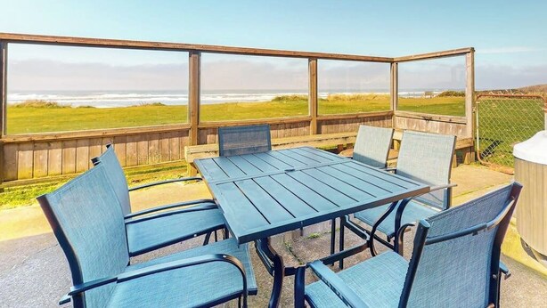 An outdoor patio with a table and chairs, overlooking a grassy area and the ocean. - Dog-friendly oceanfront beach home with private hot tub and stunning ocean views (Yachats)