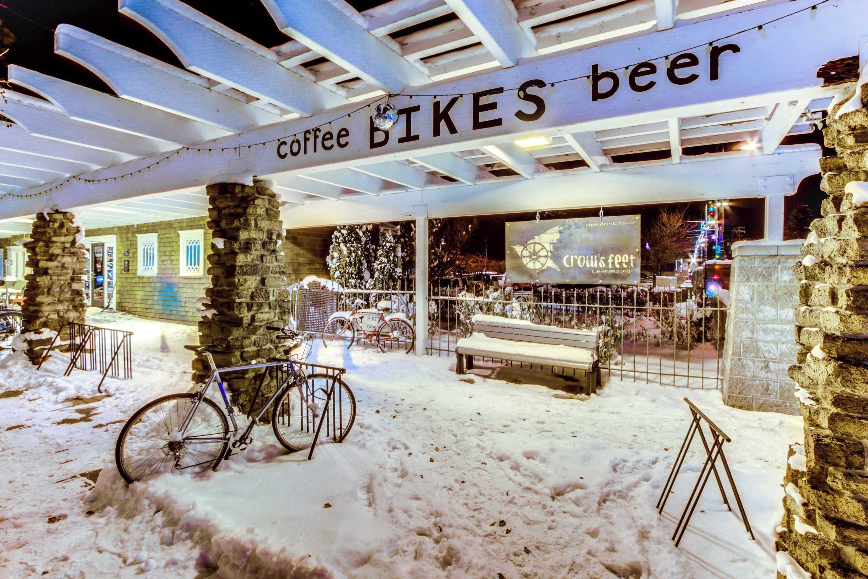 Un espace extérieur enneigé avec un vélo, un banc et un panneau indiquant : « café, VÉLOS, bière ».