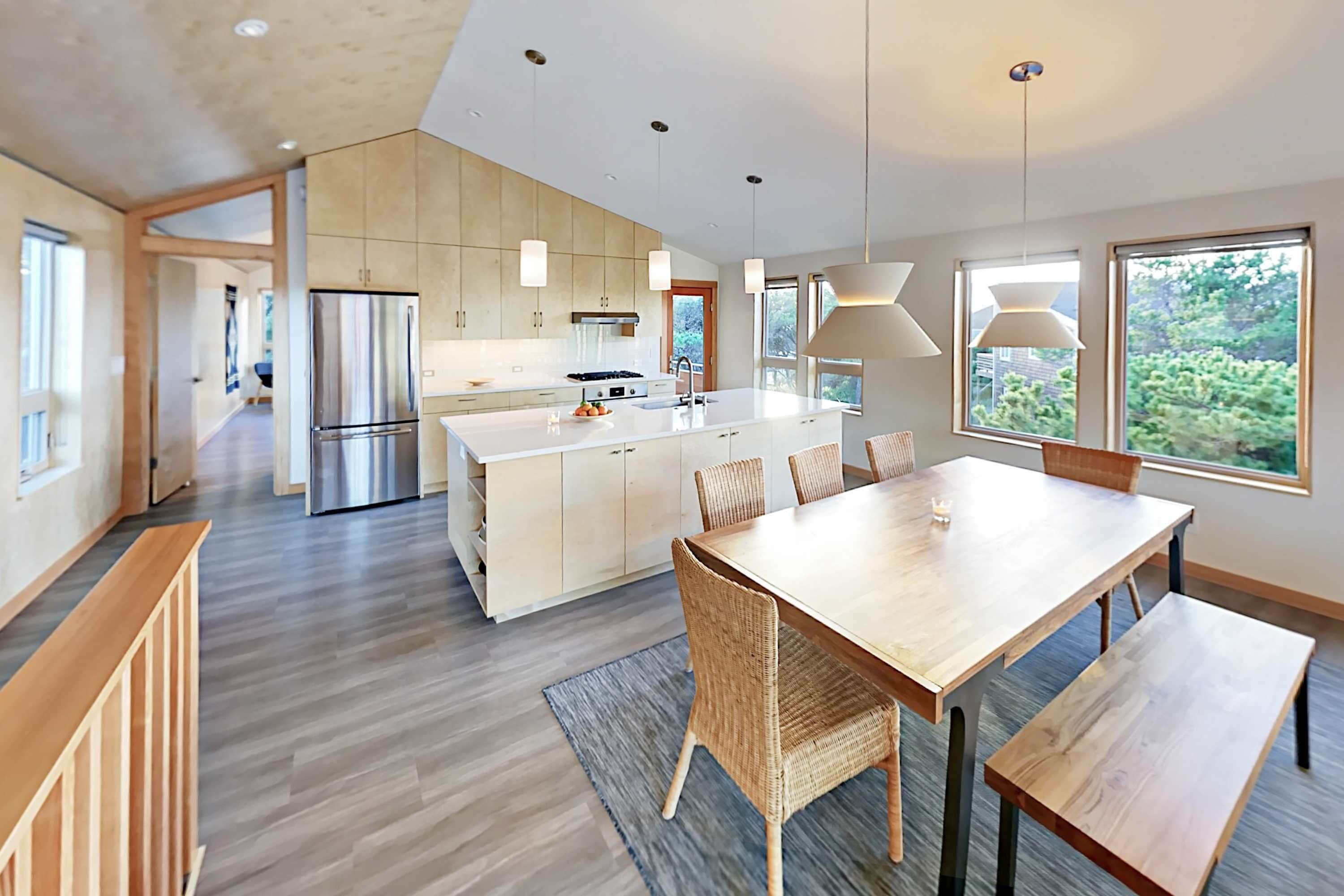 A modern kitchen with a central island, wooden dining table, and chairs, with large windows and a view of greenery.