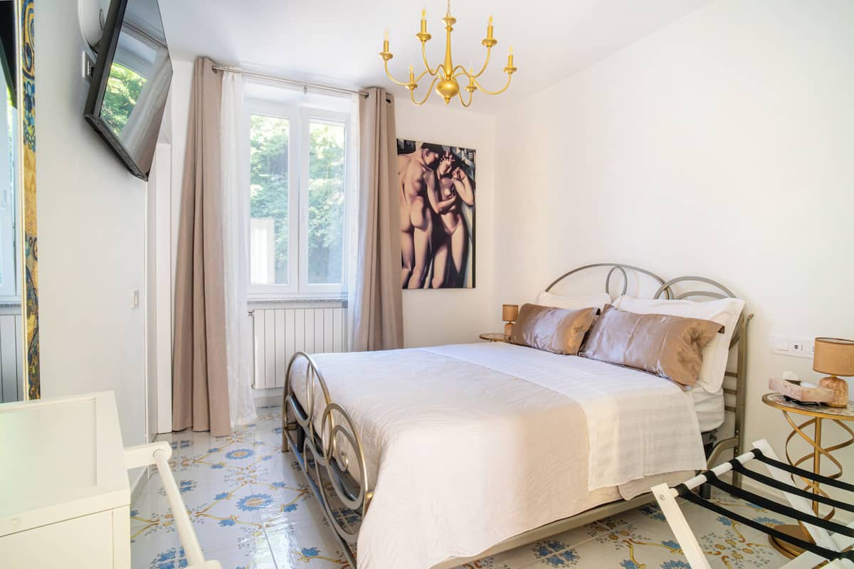 Une chambre à coucher avec un lit, un téléviseur fixé au mur, un lustre et une fenêtre avec des rideaux.