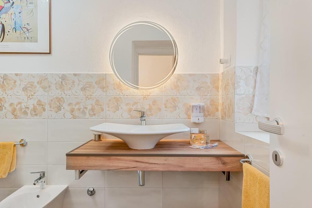 Un baño con tocador de madera, un espejo redondo con luces, un lavabo, un perchero para toallas y un cuadro enmarcado en la pared.