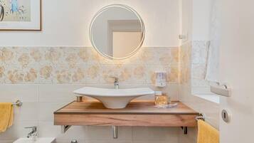Un baño con tocador de madera, un espejo redondo con luces, un lavabo, un perchero para toallas y un cuadro enmarcado en la pared.