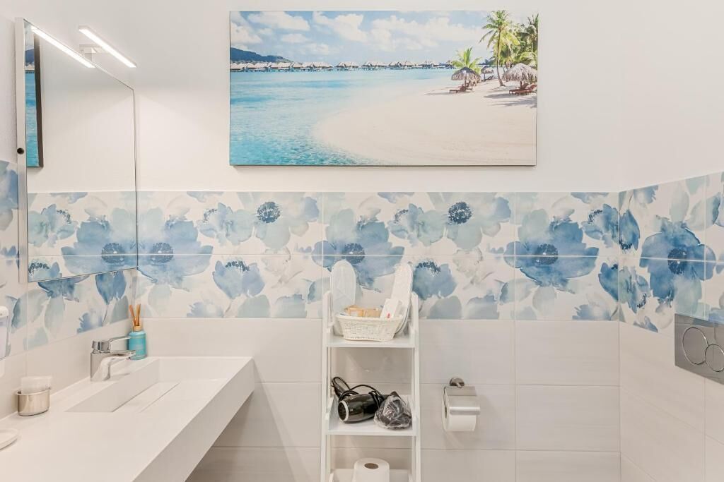 A bathroom with a white sink, a mirror, a shelf with toiletries, and a beach-themed painting.