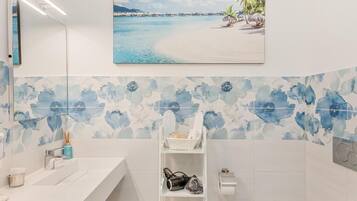 A bathroom with a white sink, a mirror, a shelf with toiletries, and a beach-themed painting.