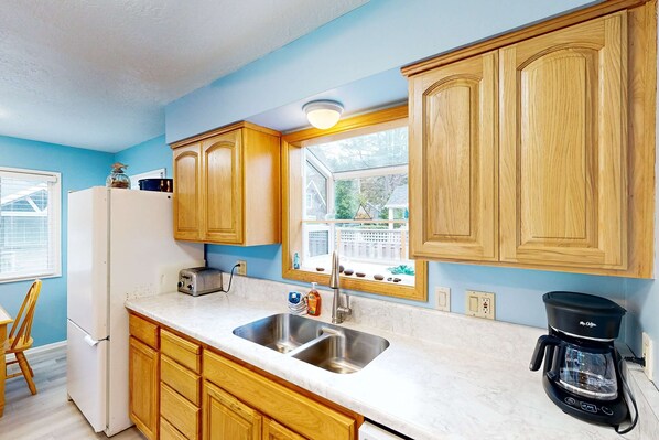 A kitchen with wooden cabinets, a white refrigerator, a double sink, a toaster, and a coffee maker. - Dog-Friendly Home Close to the Beach (Cannon Beach)