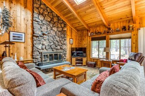 A cozy cabin interior with a stone fireplace, wooden walls, and a large window overlooking a forest. - Dog-Friendly Home with Private Hot Tub & Patio - SHARC Passes to Pools & Tennis (Sunriver)