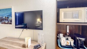 Un televisor de pantalla plana montado en una pared con textura, un microondas, una cafetera y una bandeja con varios objetos sobre una encimera de madera.