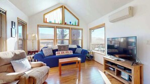 A living room with a blue sofa, beige sofa, wooden coffee table, TV stand, and large windows. - Dog-friendly cabin withprivate hot tub, large deck & beautiful ocean mountain view (Brookings)