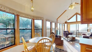 En rummelig stue med store vinduer, et spiseområde med træstole, en sofa og en loftsvifte.