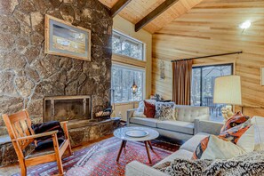 A cozy living room with a stone wall, a fireplace, a sofa, a chair, and a round table. - Dreamy mountain escape with hot tub, pools, golf, gym, & grill (Black Butte Ranch)
