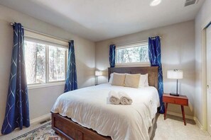 A bedroom with a large bed, two bedside lamps, and a window with curtains. - Charming home in wooded setting with deck, BBQ grill, WiFi, and fireplace (Sunriver)