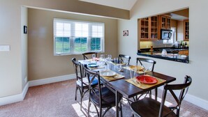 Un coin repas avec une table dressée pour six personnes, six chaises, une fenêtre avec des stores et une cuisine visible à travers une porte ouverte.