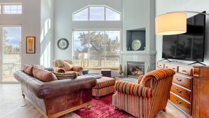 A spacious living room with a fireplace, a television, and a large window overlooking a deck. - Golf course-front home with deck, hot tub, fireplace, W&D, & community pool (Eagle Crest)