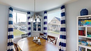 A dining area with a wooden table, chairs, and a chandelier. There is a window with curtains, a bookshelf with books, and a view of houses outside. - Dog-Friendly Home w/ Gas Fireplace & Patio, in a Great Location Near the Beach (Newport)