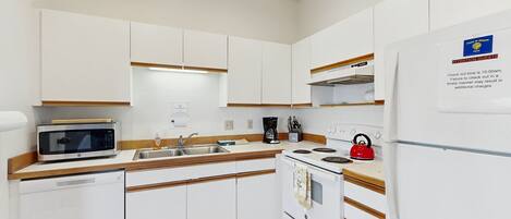 Una cocina con armarios blancos, microondas, cafetera, hervidor y refrigerador.