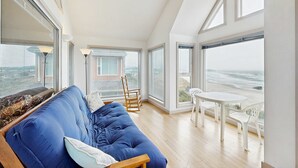 Een kamer met een blauwe bank, een houten tafel en stoelen, grote ramen met uitzicht op zee, en een hoog plafond.