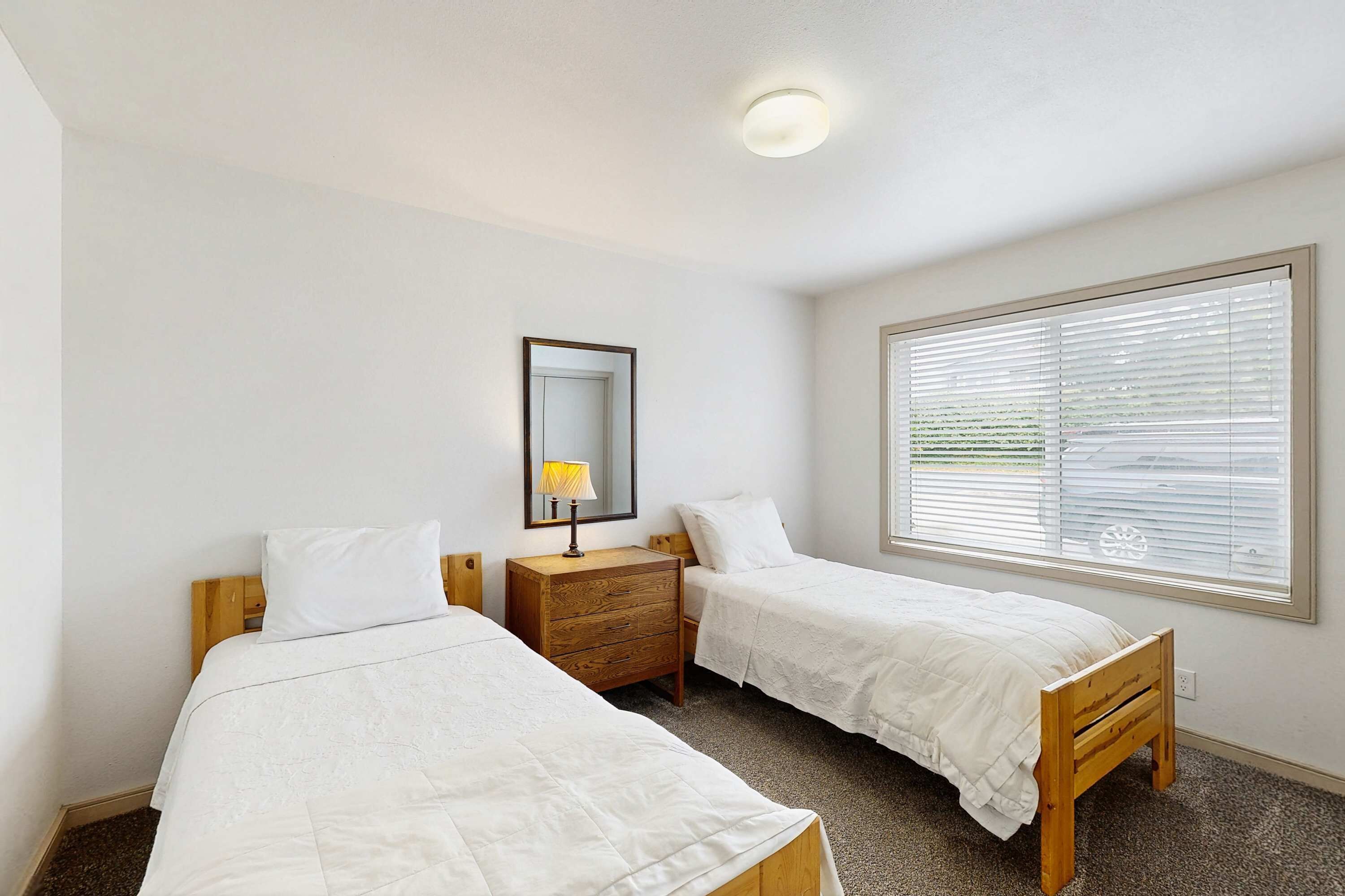 Ein Schlafzimmer mit zwei Einzelbetten, einer Holzkommode, einem Spiegel und einem Fenster mit Jalousien.