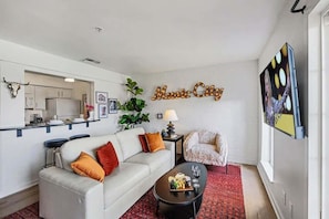 Una sala de estar con un sofá, una mesa de centro y un televisor. Se ve una cocina a través de un acceso abierto.