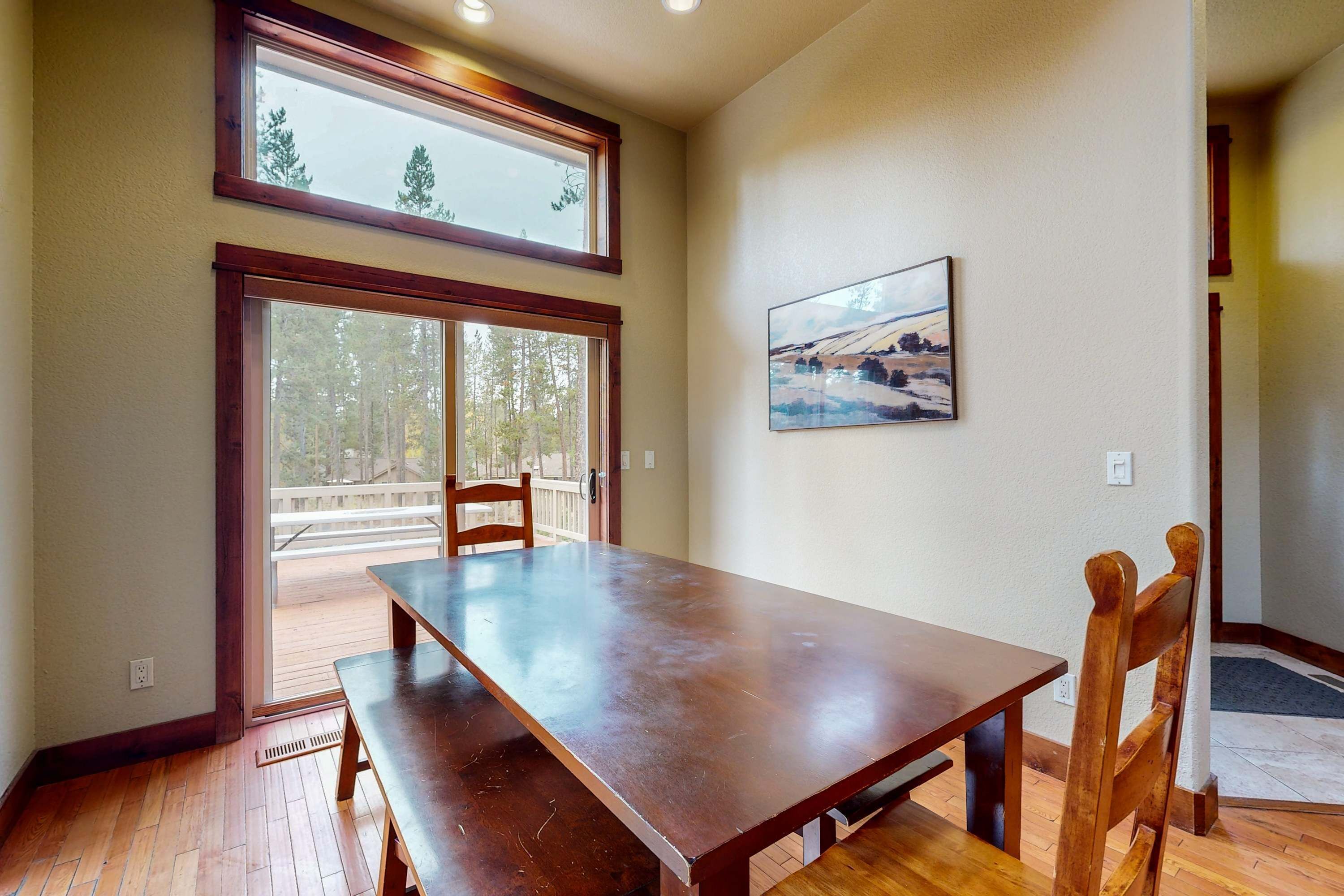 A dining area with a wooden table and chairs, a large window with a view of trees, and a painting on the wall.