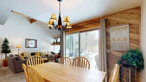 A dining area with a wooden table and chairs, a chandelier, and a large window with curtains. - Open & modern condo with pool, hot tub, fireplace & W&D (Sunriver)