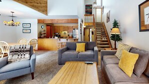 A spacious living area with a sofa, armchairs, and a coffee table. A kitchen is visible in the background, and a staircase leads to an upper level. - Open & modern condo with pool, hot tub, fireplace & W&D (Sunriver)