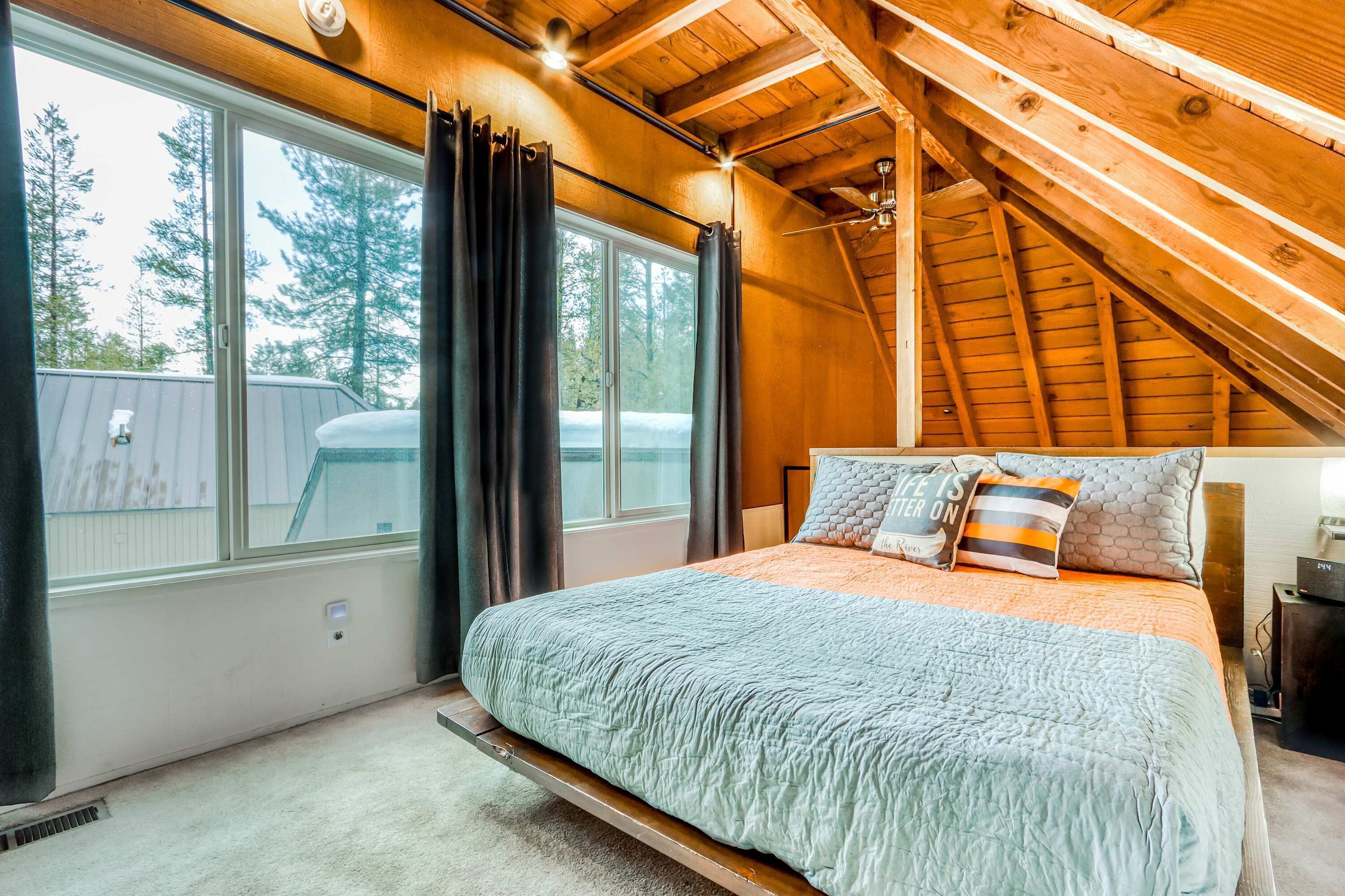 Un dormitorio con techo inclinado, una cama grande con colcha gris, dos almohadas y una ventana con cortinas.