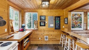 A cabin interior with wooden walls, a kitchen area with a stove, a bar with stools, and framed pictures on the walls.